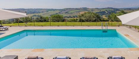 Une piscine rectangulaire à l’eau d’un bleu cristallin, entourée de chaises longues et offrant une vue sur des collines verdoyantes à l’arrière-plan.
