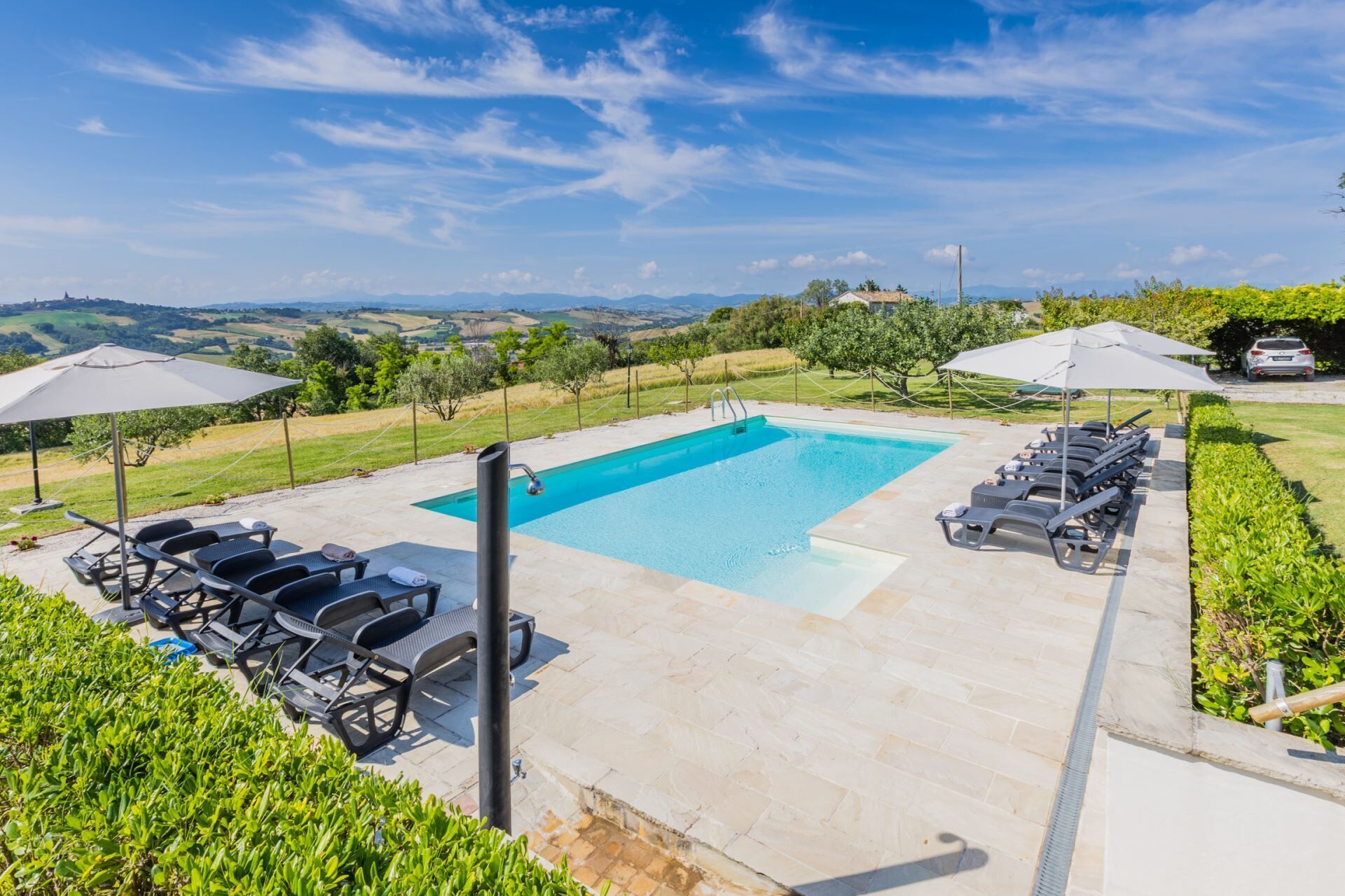 A rectangular swimming pool with a clear blue water surface, surrounded by a paved deck. There are lounge chairs and umbrellas arranged around the pool, and a view of the countryside in the background.