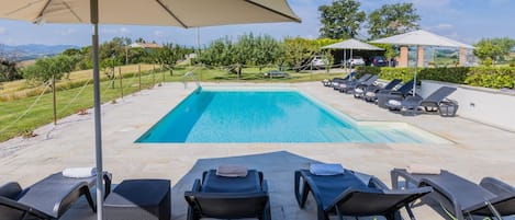 A rectangular swimming pool with a clear blue water, surrounded by lounge chairs and umbrellas, set on a stone patio with a view of the countryside.