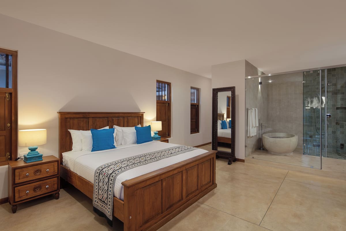 A bedroom with a wooden bed, a nightstand, a bedside table, a large mirror, and a glass-enclosed shower area.