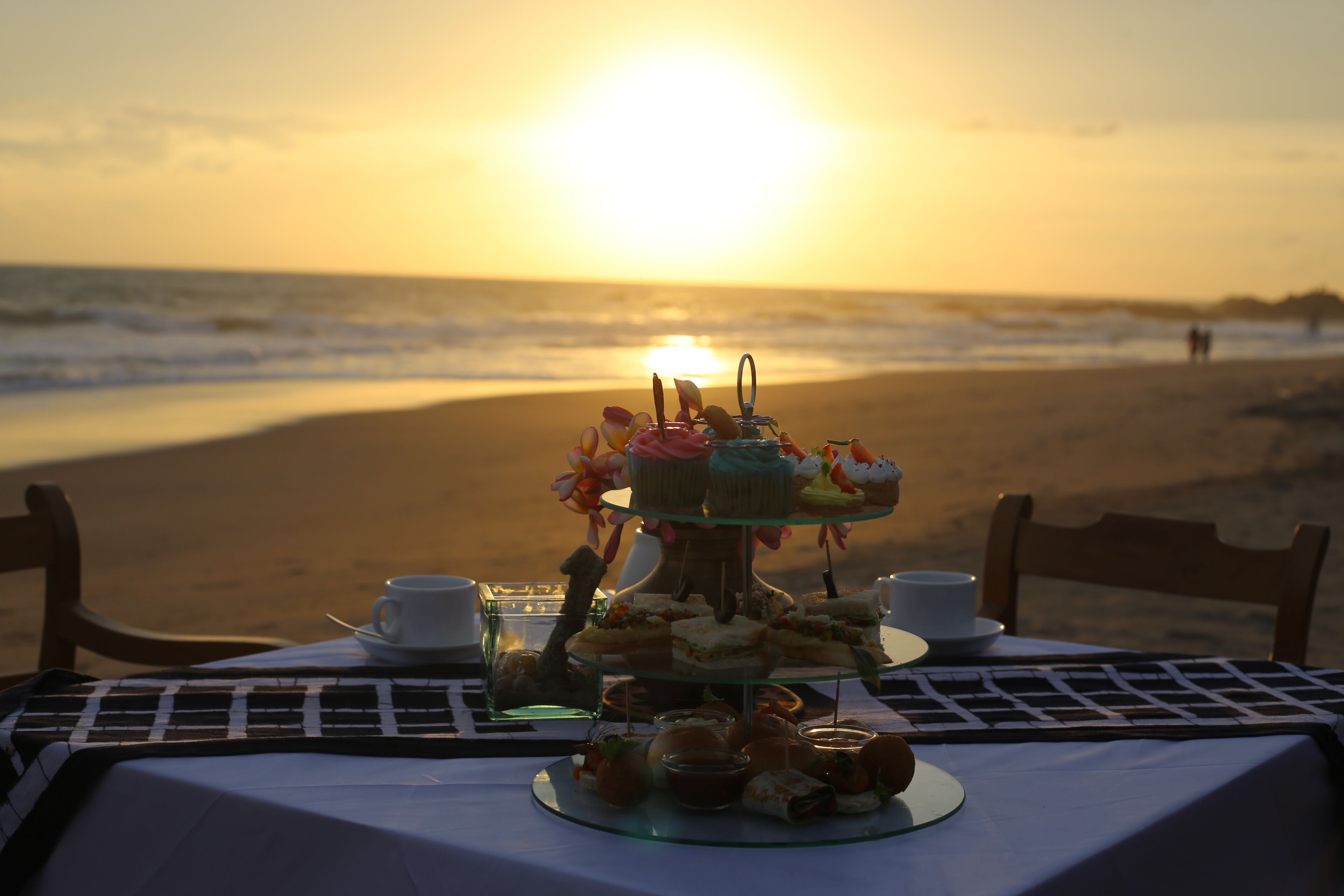 A table set with a variety of pastries and sandwiches on a beach at sunset.