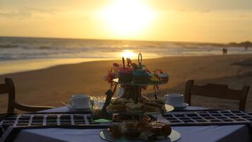 A table set with a variety of pastries and sandwiches on a beach at sunset.