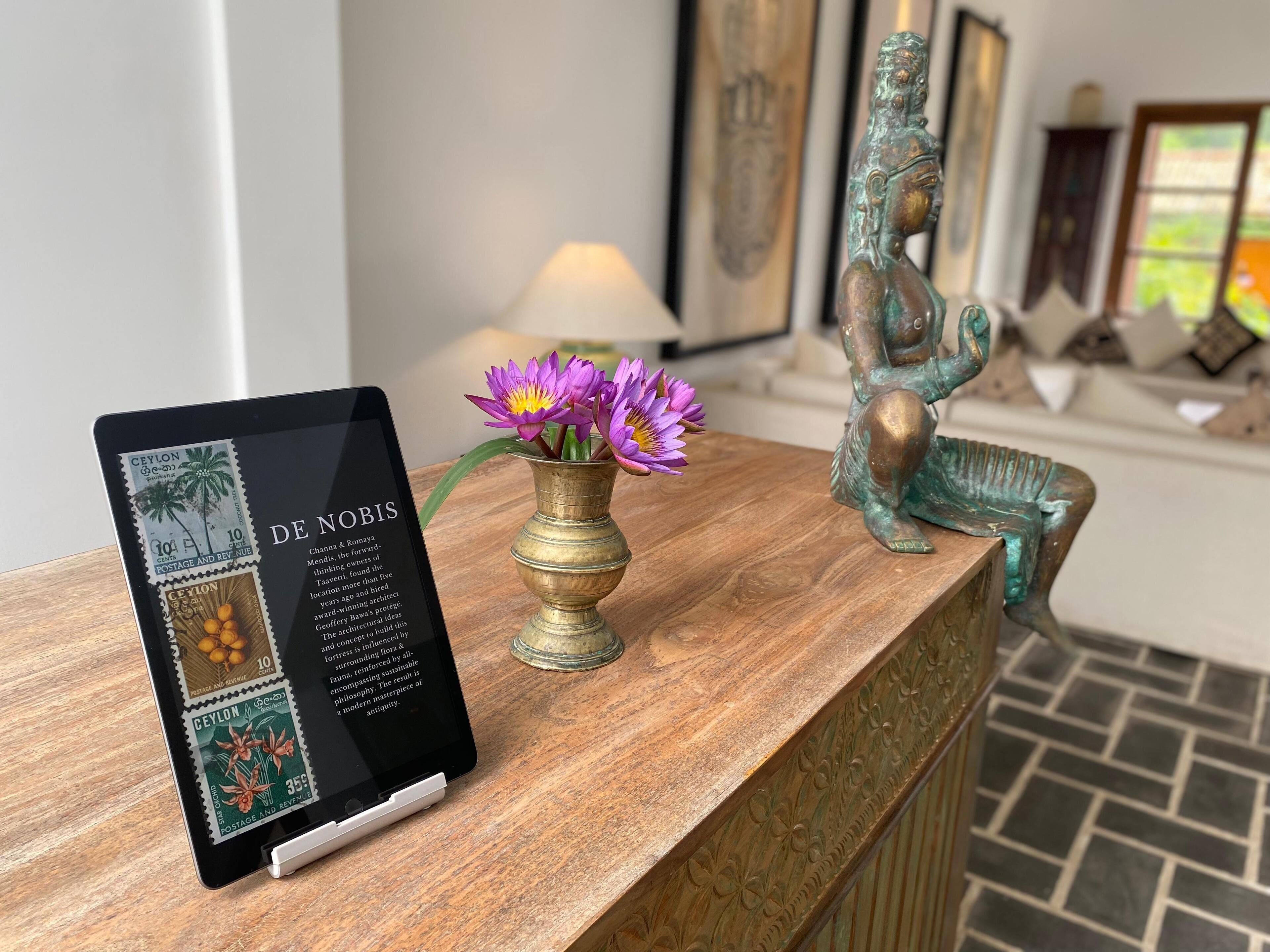 Un tablet mostra informazioni con la scritta 'DE NOBIS' su una superficie in legno, un vaso con fiori viola e una statua in bronzo.