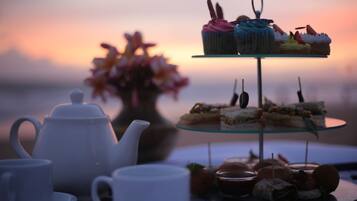 A tea set, a tiered dessert stand with various pastries, and a vase of flowers on a table with a sunset view in the background.