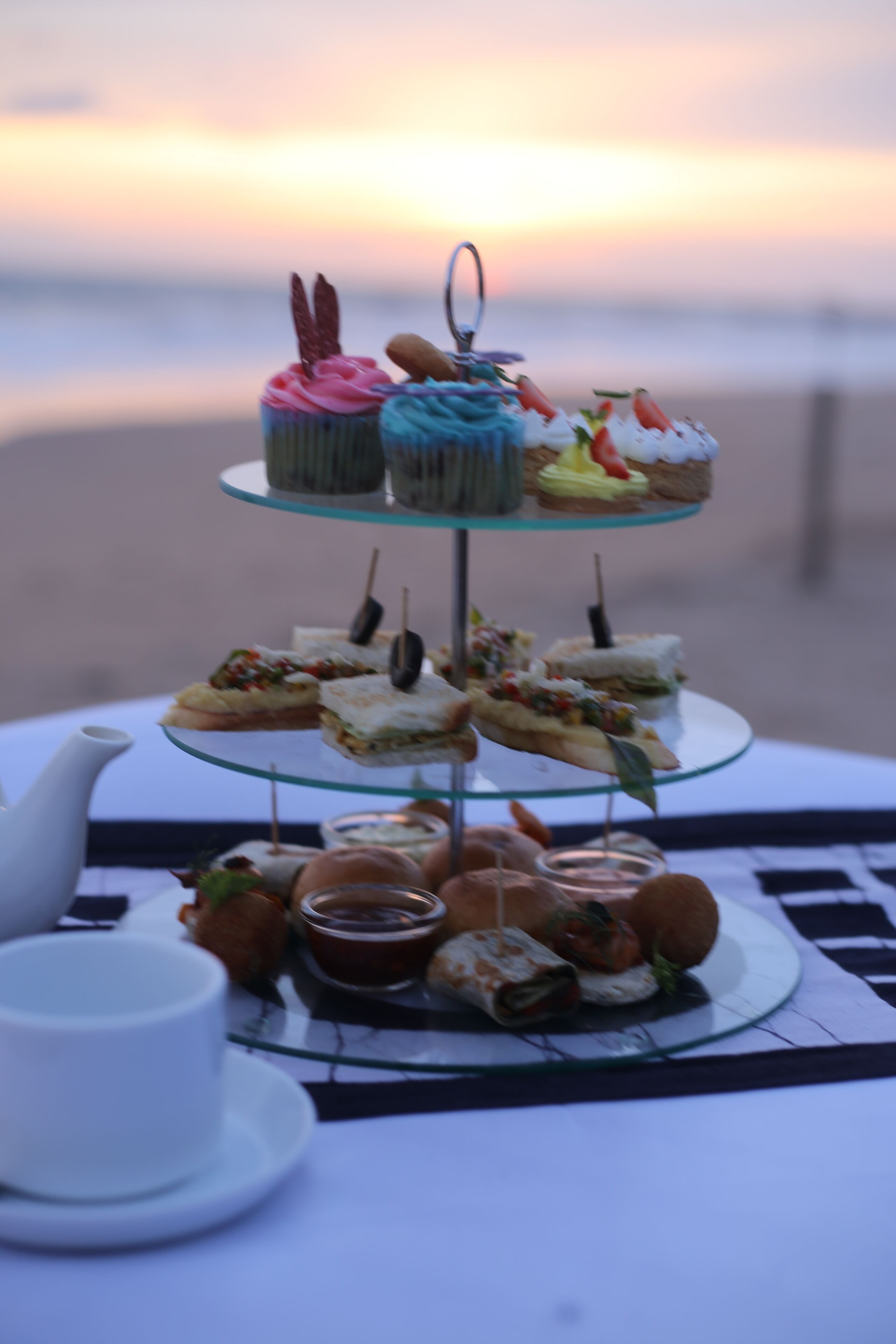 Un présentoir à gâteaux en verre à trois niveaux, garni de diverses pâtisseries et de sandwichs, accompagné de tasses et d’une théière, est placé sur une table offrant une vue sur la plage au coucher du soleil.