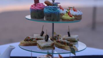 Un présentoir à gâteaux en verre à trois niveaux, garni de diverses pâtisseries et de sandwichs, accompagné de tasses et d’une théière, est placé sur une table offrant une vue sur la plage au coucher du soleil.