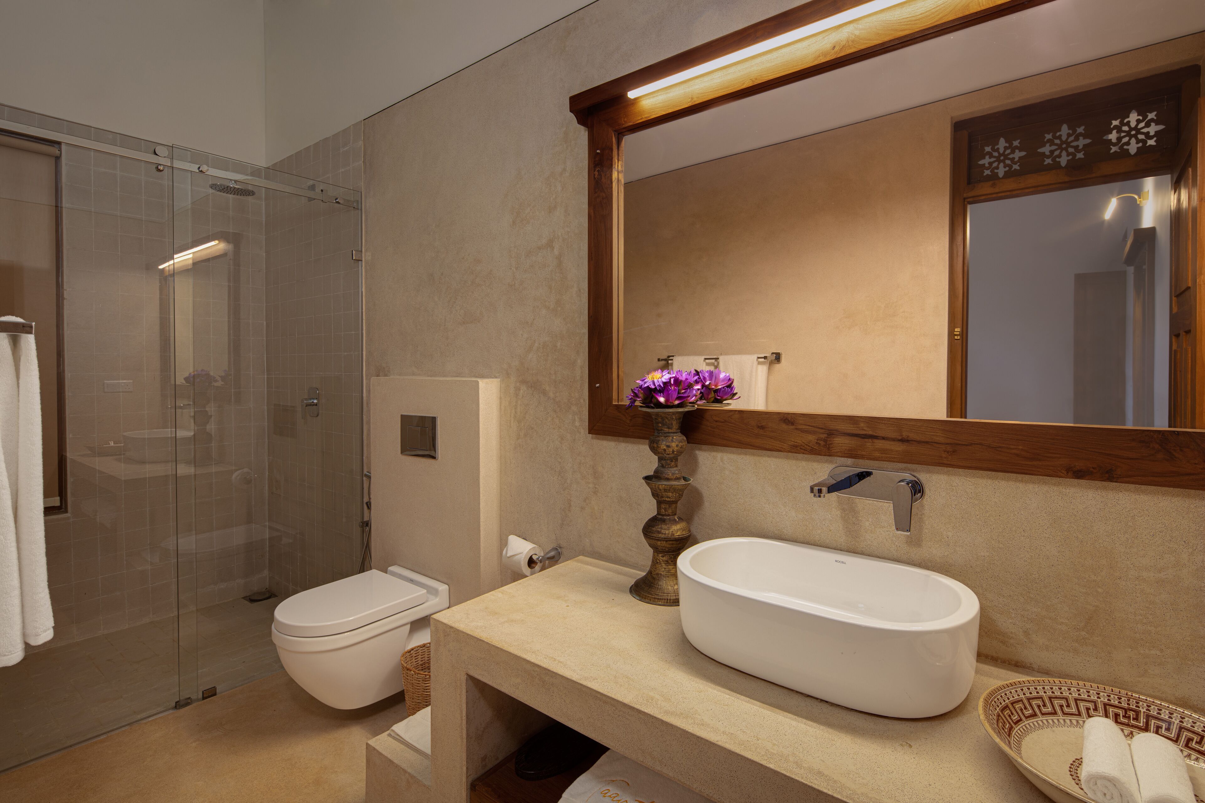 Un baño moderno con cabina de ducha de vidrio, inodoro blanco, lavabo color beige y un espejo grande, además de un jarrón decorativo con flores moradas.
