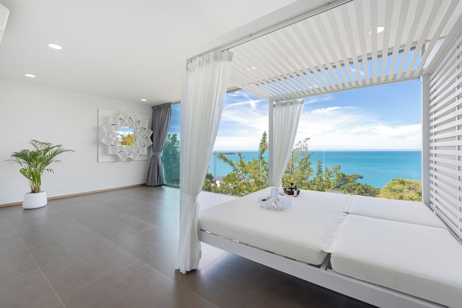 Une chambre moderne dotée d’une grande fenêtre donnant sur un magnifique paysage marin, d’un lit blanc et d’une plante en pot.