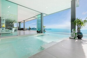 Une piscine à débordement moderne offrant une vue dégagée sur l’océan, un salon vitré et une plante en pot.