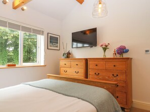 A bedroom with a large bed, wooden dresser, a flat-screen TV, and a window with a view of trees. - Blake Cottage (Looe)