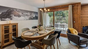 A dining area with a wooden table set for four, surrounded by modern chairs, a wooden cabinet, and a large mountain painting on the wall.