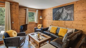 A cozy living room with wooden walls, a black leather sofa, a glass coffee table, and a mountain landscape painting.
