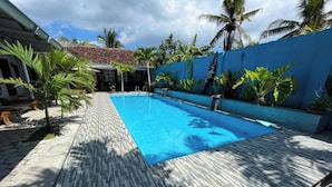 A swimming pool with a tiled deck, surrounded by palm trees and a blue wall. - Capital O 92615 Villa Utama D'alas Purwo (Kendalrejo)