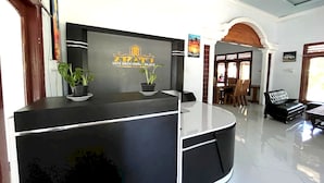 A reception desk with a logo, two potted plants, and a dining area with chairs and a table in the background. - Capital O 92615 Villa Utama D'alas Purwo (Kendalrejo)