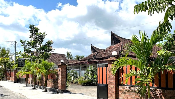 A row of traditional-style buildings with tiled roofs and brick walls, surrounded by palm trees and a paved walkway. - Capital O 92615 Villa Utama D'alas Purwo (Kendalrejo)