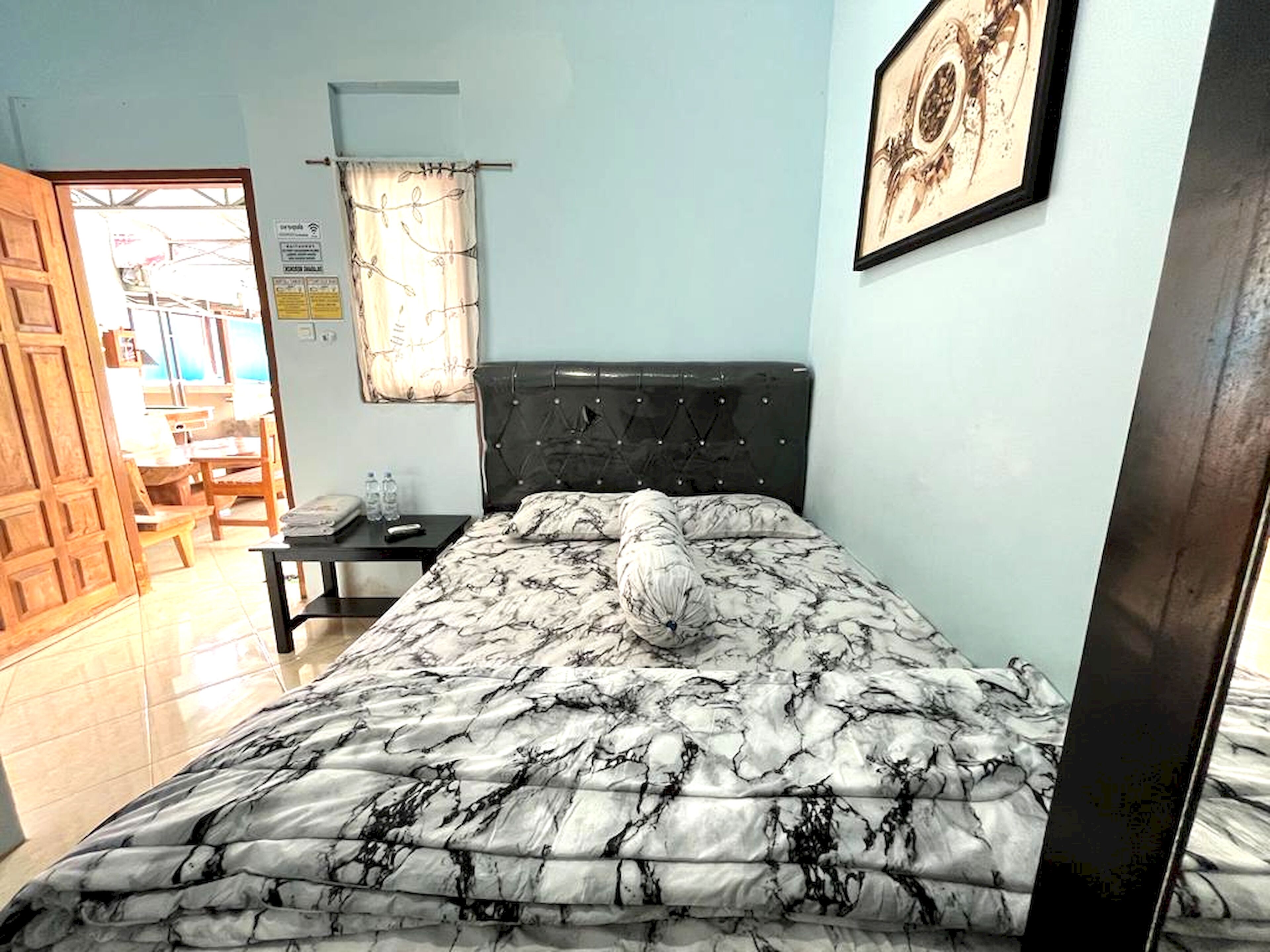 Quarto com cama, mesa de cabeceira, quadro na parede e porta que dá para outro cômodo.