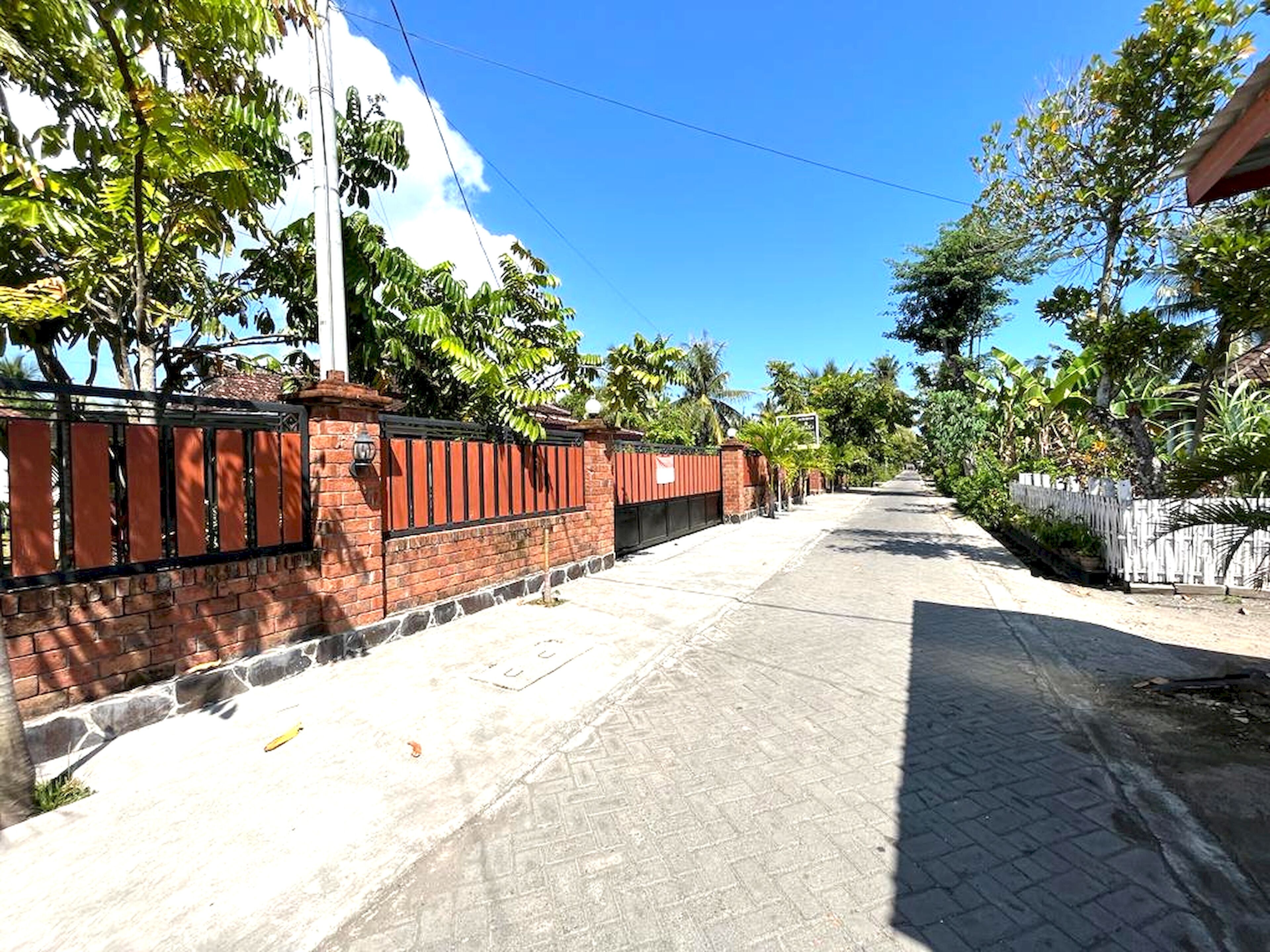 Rua residencial tranquila com muro de tijolos e portão de metal, rodeada por vegetação exuberante e céu azul límpido.