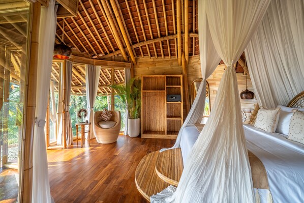 A spacious room with a wooden floor, a bed with white linens, a wooden wardrobe, and a large window offering a view of greenery. - Bambootel Sawah View (Tampaksiring)