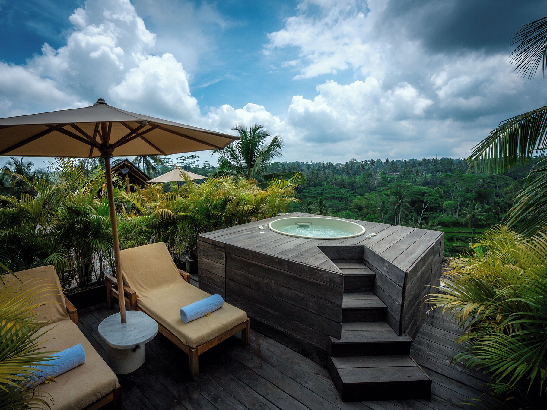 A wooden hot tub with steps leading up to it, surrounded by lounge chairs and umbrellas, overlooking a lush landscape.