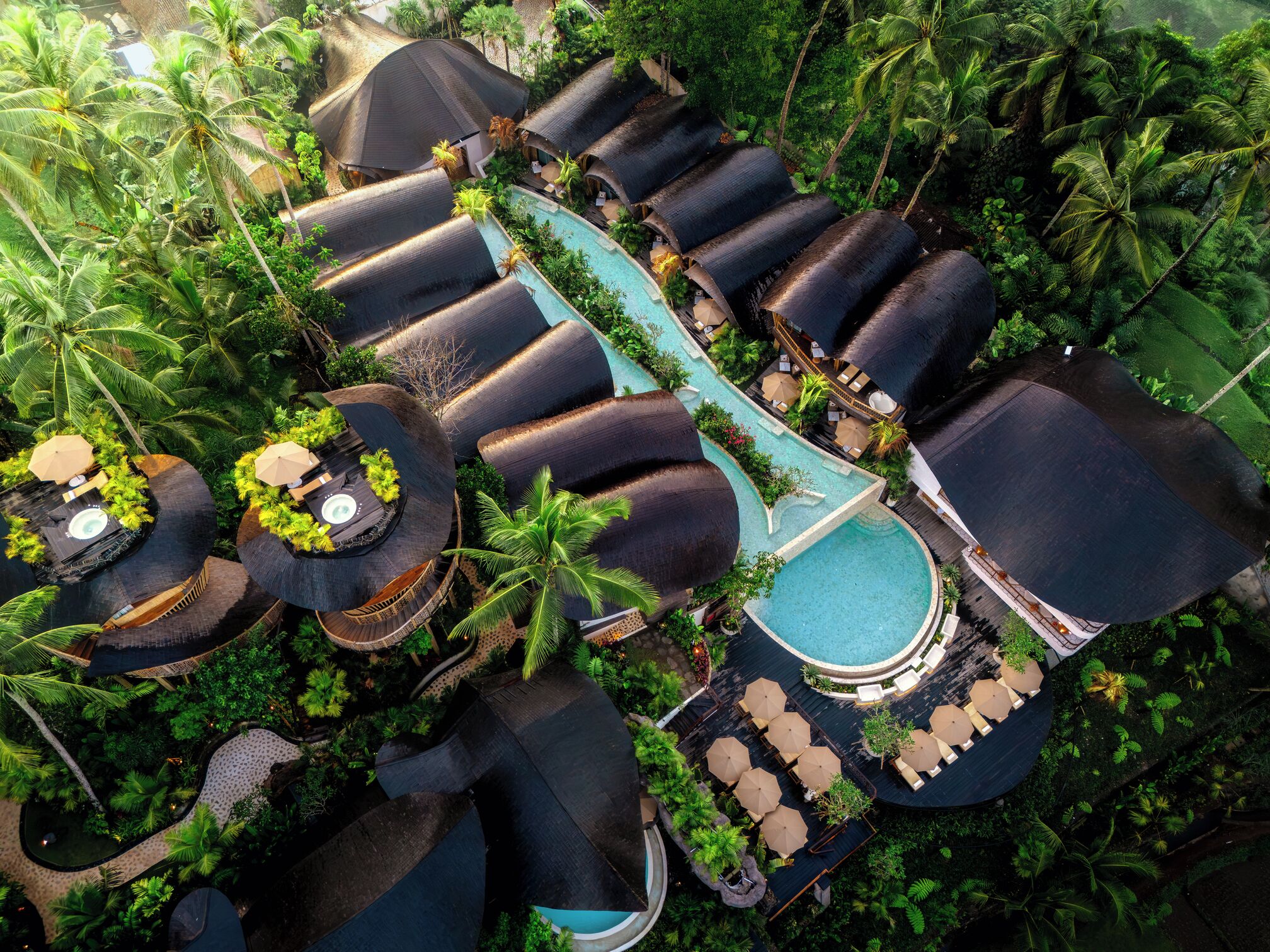 An aerial view of a resort with multiple overwater bungalows, a central pool, and lush tropical vegetation.