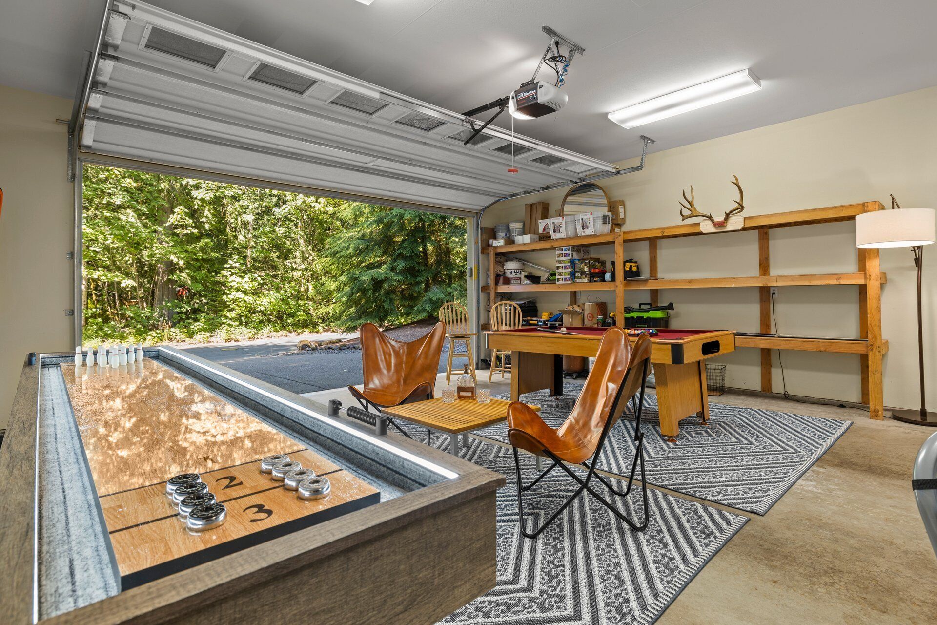 A room with a billiards table, chairs, a wooden desk, shelves with various items, and a view of greenery outside.