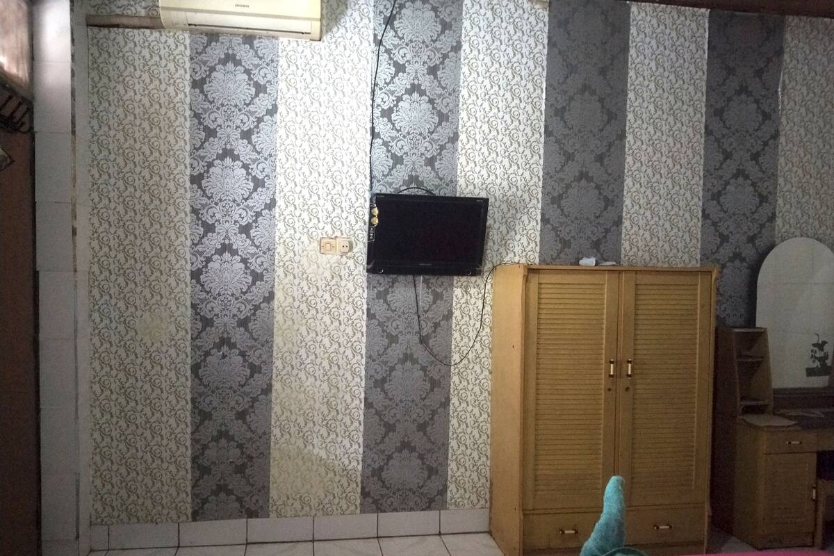 A bedroom with patterned wallpaper, a wooden wardrobe, a television mounted on the wall, and a pink bedspread.