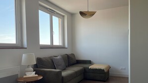 A modern living room with a sectional sofa, a small round table with a lamp, a throw blanket, and a large window providing natural light. - The Lighthouse (Calais)