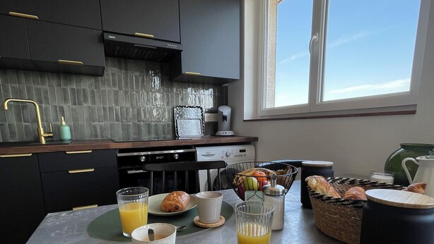 A modern kitchen with dark cabinetry, a large window, and breakfast items on the counter. - The Lighthouse (Calais)
