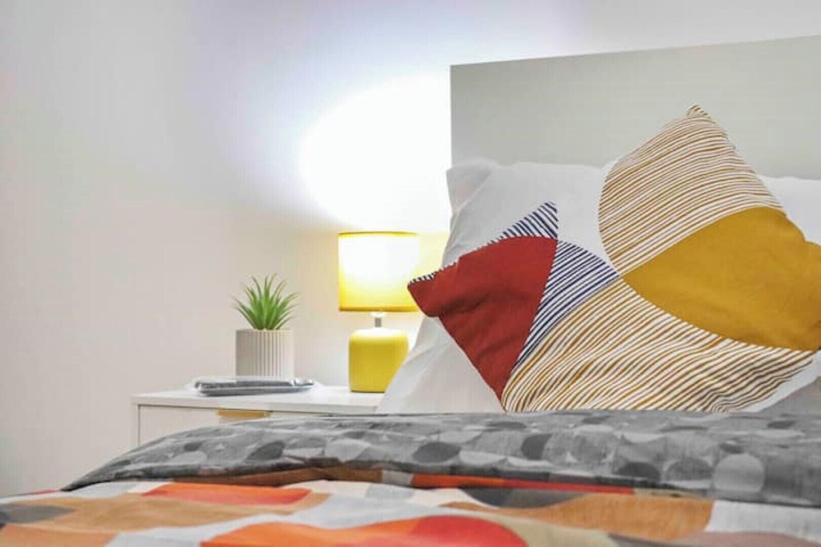 Un lit bien fait, avec une couverture à motifs, une lampe de chevet, une petite plante en pot et plusieurs coussins de différentes sortes.