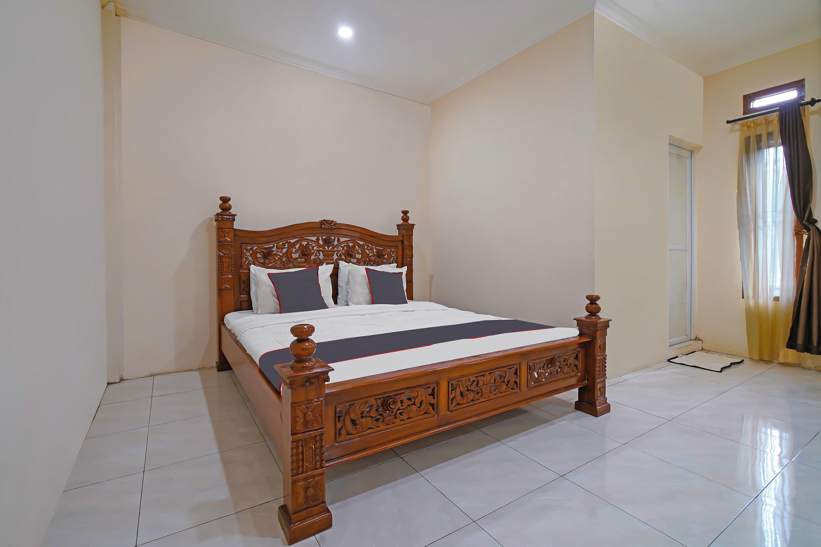 A neatly made bed with a wooden frame and a white bedspread, a window with curtains, and a tiled floor.