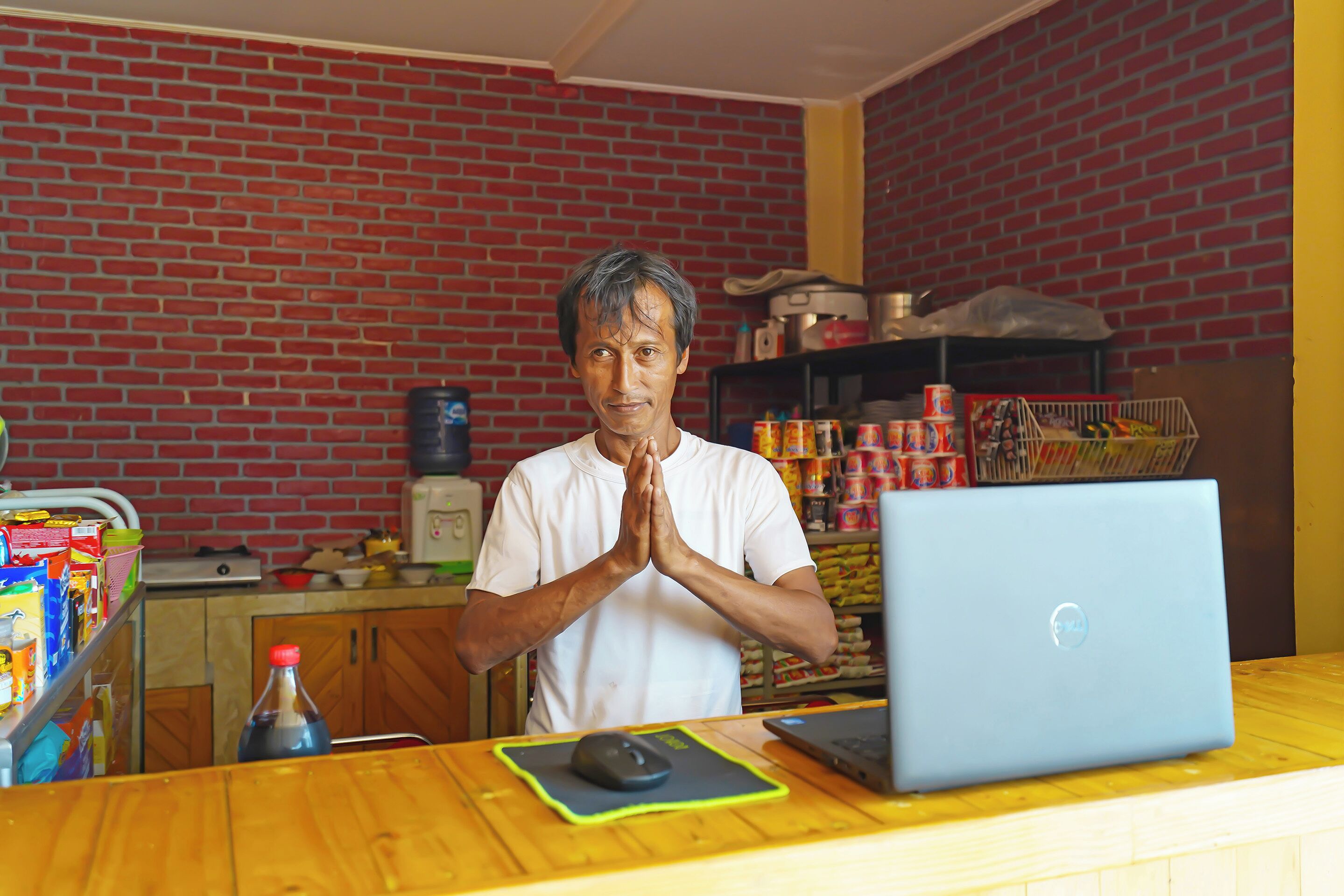 A person standing in a room with a laptop on a wooden desk, a brick wall, and shelves with various items.