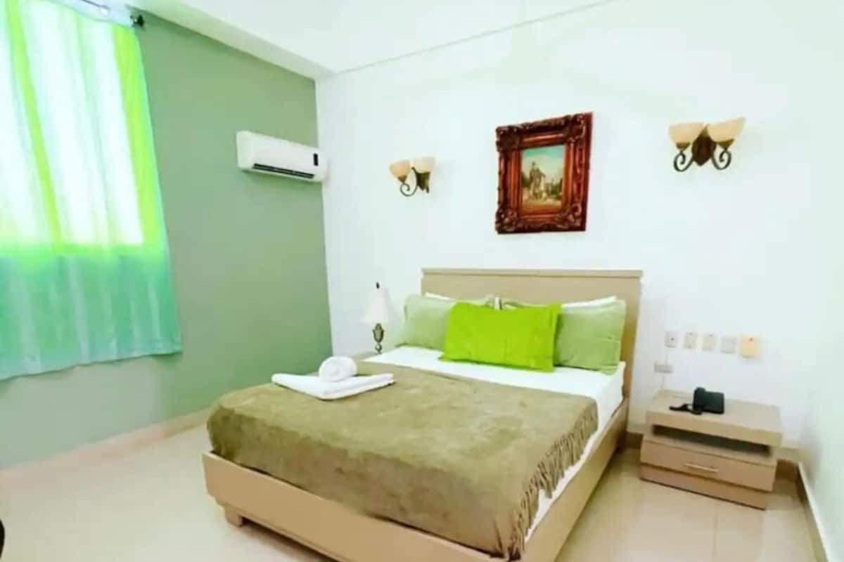 A bedroom with a bed, bedside table, wall-mounted air conditioner, and a picture frame.