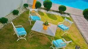 Un patio con mesa y cuatro sillas, rodeado de un césped pequeño y una piscina.