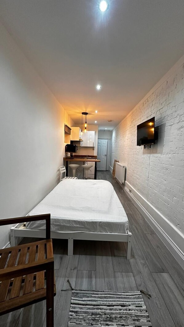 A compact room with a bed, a wooden bench, a TV mounted on the wall, and a kitchenette in the background.