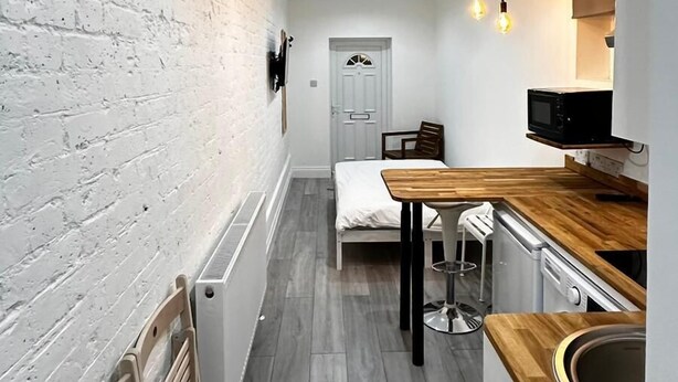 A narrow hallway with a white brick wall, a wooden floor, a bed, a small table, and a microwave. - Spacious 3 Bed Flat in Greenwich for 6 (London)
