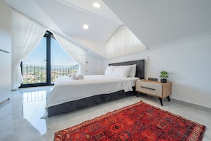 A modern bedroom with a large bed, a nightstand, a red patterned rug, and a view of a cityscape through a balcony.