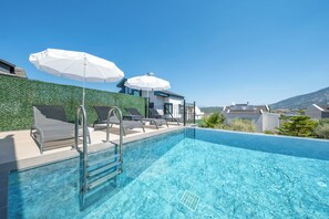 Una piscina azul cristalina con sillas de descanso y sombrillas, un edificio blanco con un muro verde, y vistas del paisaje.