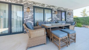 Un patio al aire libre con mobiliario de mimbre, paredes de piedra y grandes ventanales.