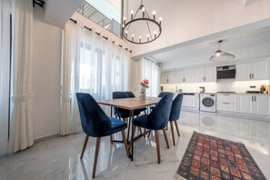Una cocina moderna con armarios blancos, un comedor con mesa de madera y sillas azules, y una lámpara de araña.