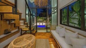 Un interior moderno con una escalera de madera, una zona de estar con cojines y vistas a una piscina.