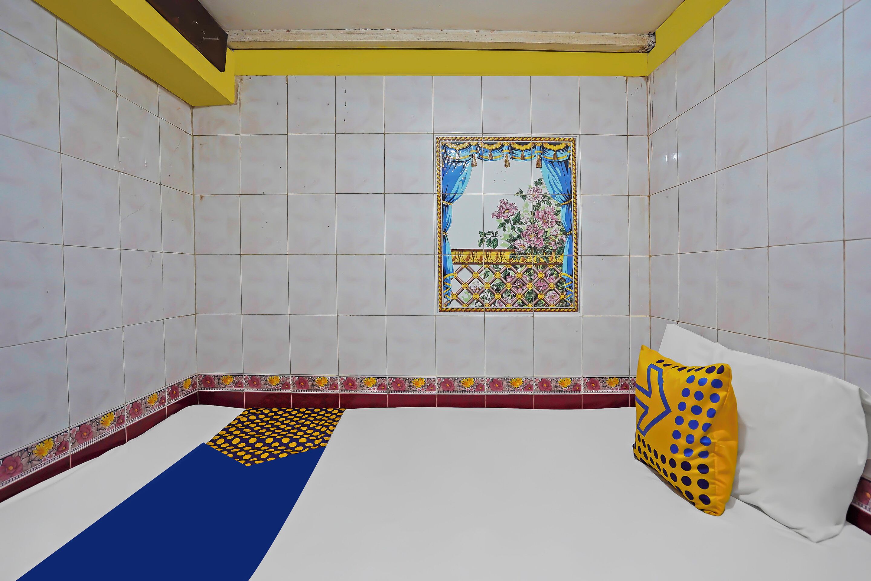 A room with white tiled walls, a decorative window, a bed with a blue and yellow patterned runner, and a white pillow with blue dots.