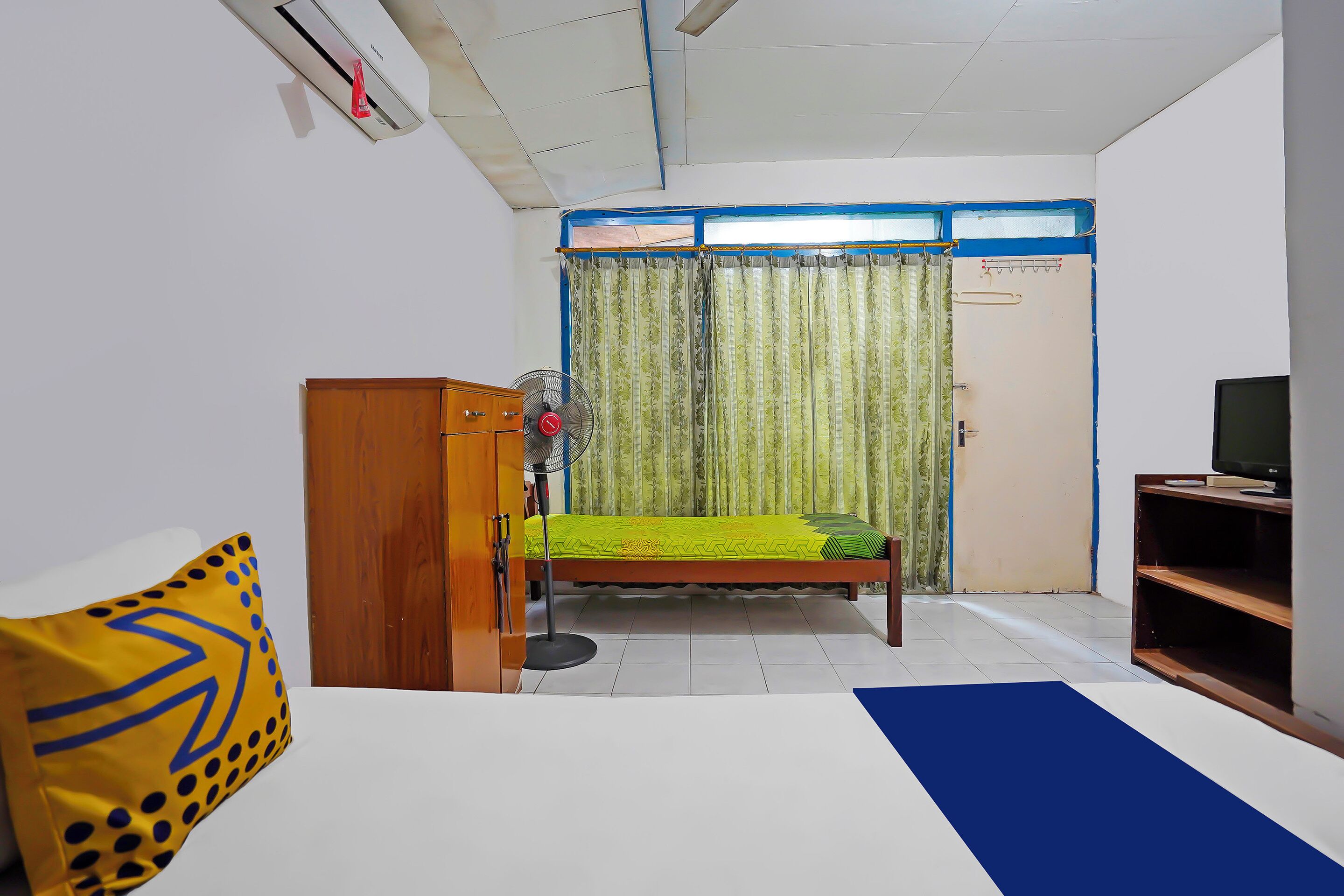 A bedroom with a bed, a wooden wardrobe, a desk with a computer, a fan, and a window with curtains.