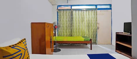 A bedroom with a bed, a wooden wardrobe, a desk with a computer, a fan, and a window with curtains.