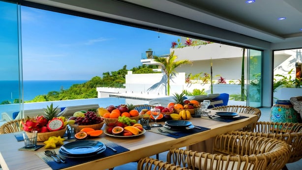 Una mesa de comedor con frutas y vasos varios, con vista a un paisaje marino.