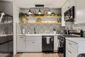 Ett modernt kök med vita skåp, ett kylskåp i rostfritt stål, en diskmaskin, en spis och en mikrovågsugn. Ovanför diskhon hänger två hänglampor, och det finns en hylla i trä med växter och köksredskap.