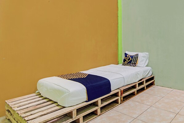 A single bed with a white mattress and blue bedding, placed on a wooden pallet base, against a yellow and green wall.