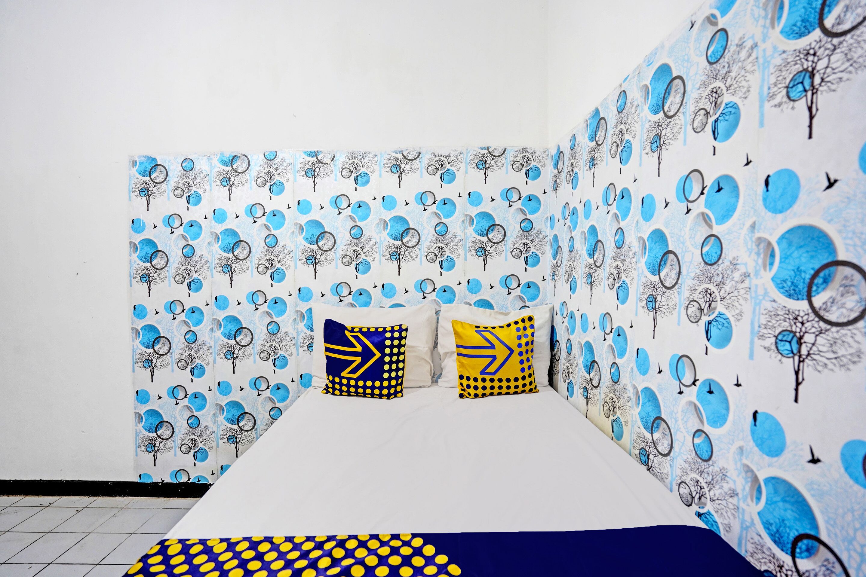 A room with a patterned wallpaper, two pillows with arrow designs, and a bed with a blue and yellow polka dot bedspread.