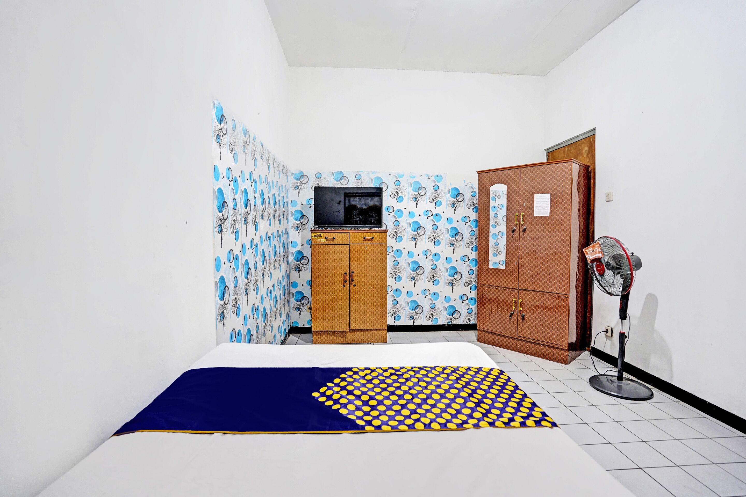 A small, sparsely furnished room with a bed, a television, a wardrobe, and a standing fan.