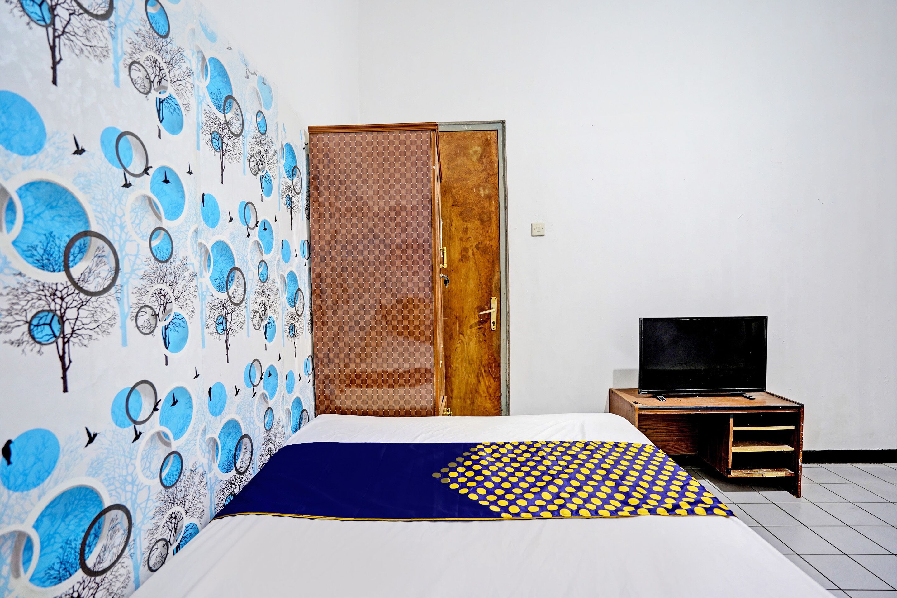 A bedroom with a bed, a TV on a wooden stand, a wardrobe, and a wall with a blue and white abstract design.
