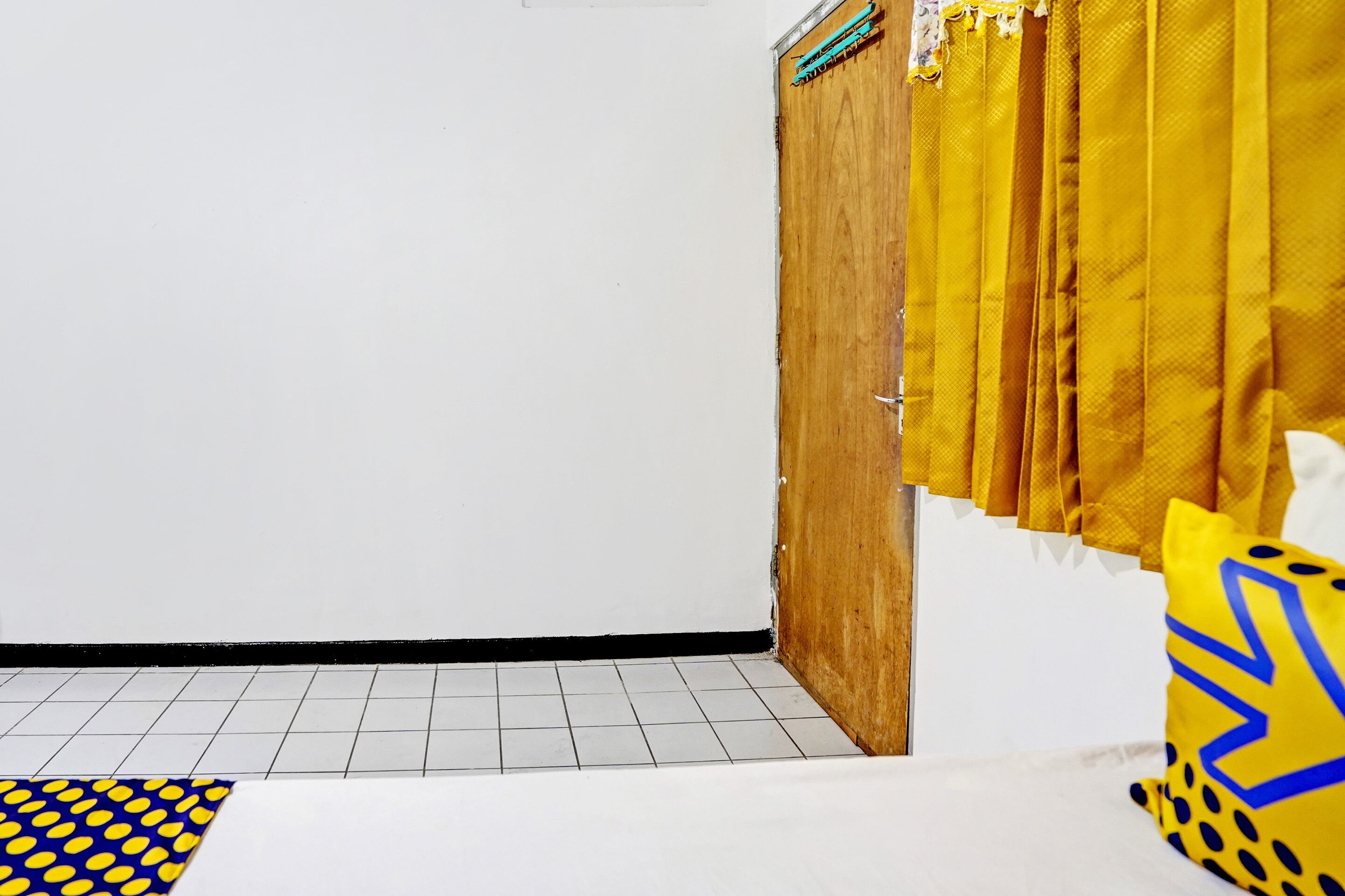 A room with a wooden wardrobe, yellow curtains, a patterned pillow, and a tiled floor.